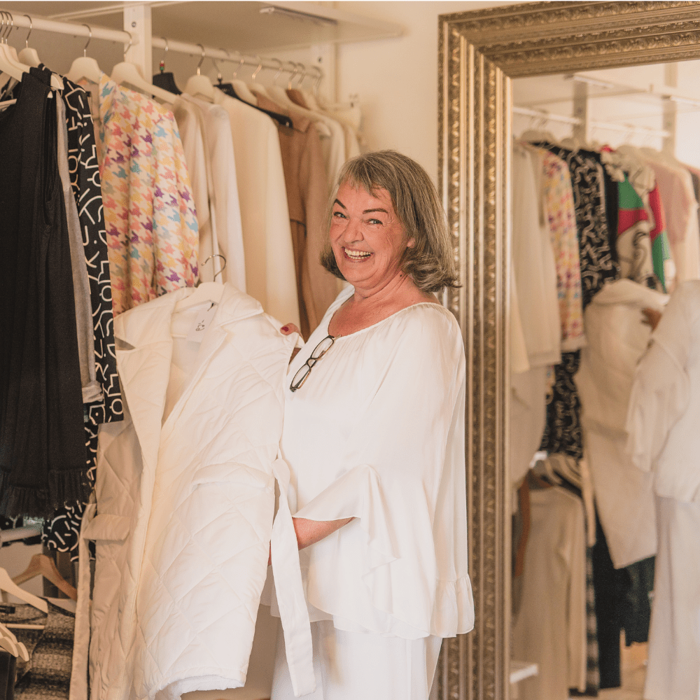Simone Klinge-Otto steht vor einem Kleiderschrank und präsentiert eine weiße Weste. Sie lacht dabei fröhlich in die Kamera. Das Bild demonstriert, dass Sie als Expertin Frauen ab 50 zu einem eigenen individuellen Stil berät.