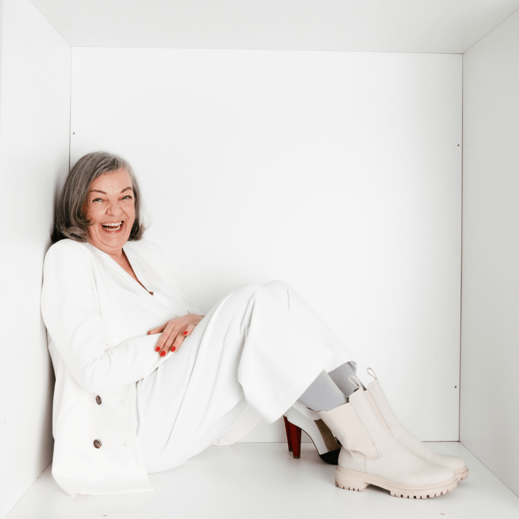 Simone Klinge-Otto sitzt mit einem weißen Hosen Anzug angelegt an einer weißen Box und lacht lebensfroh und herzlich in die Kamera.