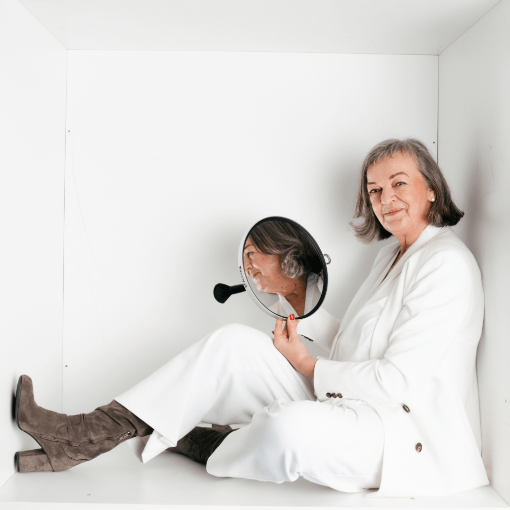 Simone Klinge-Otto bildet Friseure zu Stylecoaches aus. Sie sitzt auf dem Foto mit einem weißen Hosenanzug, hält einen Spiegel in die Hand und schaut freundlich in die Kamera.